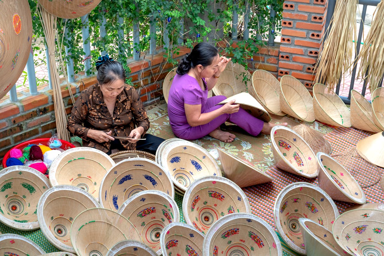 Two artisans meticulously crafting traditional Vietnamese conical hats with colorful decorations outdoors.